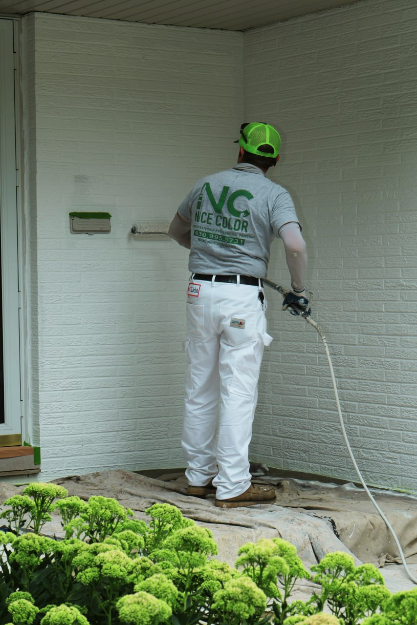 A Nice Color Painting painter in a branded gray shirt using a sprayer to paint a white brick wall of a house, with green hydrangea bushes in the foreground